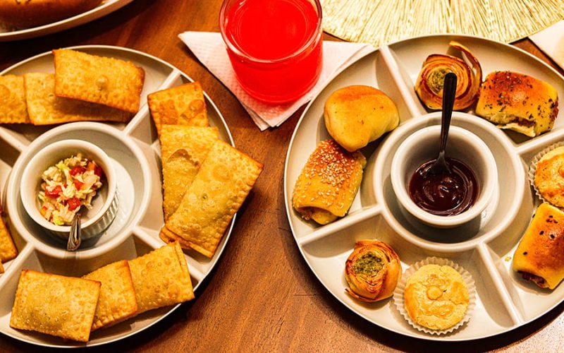 Fotografia vista de cima de duas petisqueiras redondas com salgados de festa. Uma contém minipastéis fritos com vinagrete, e a outra exibe salgados assados variados com um molho escuro ao centro. Ao fundo, um copo de suco e um cachorro-quente. Destaque para a gastronomia de um buffet em Caraguatatuba.